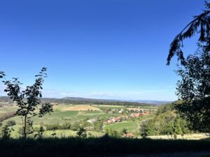 Weitblick, hügelige Landschaft mit Ackerland, Wald und Dörfern, darüber Blauhimmel