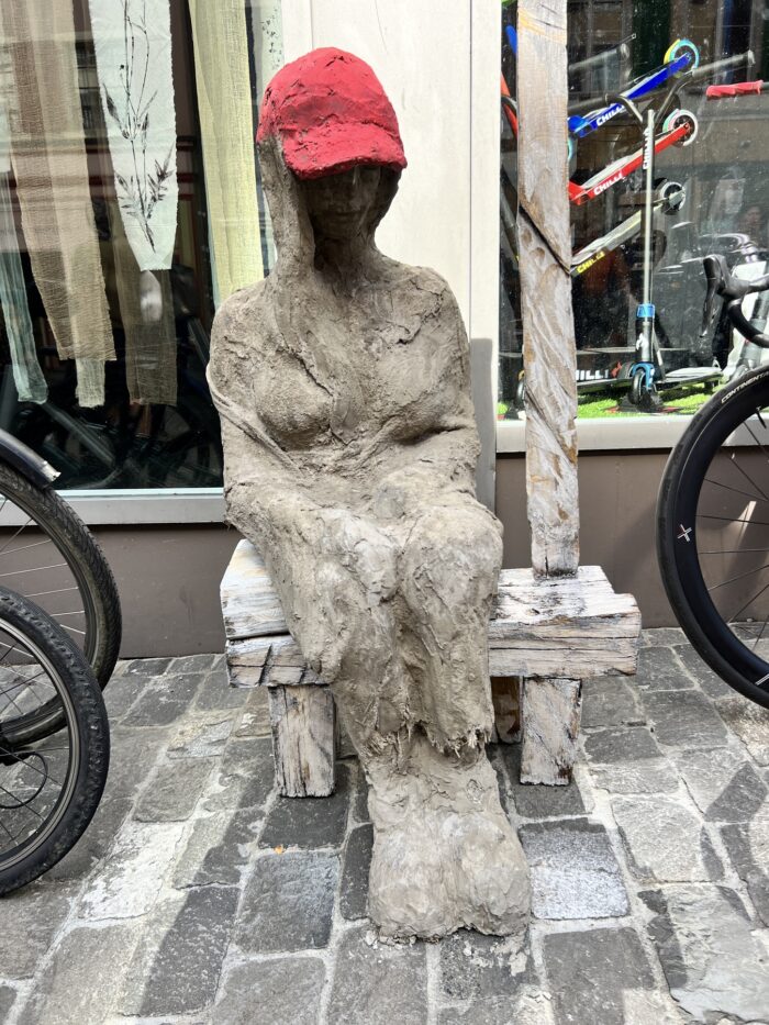menschliche Skultpur aus Ton oder Beton, die auf einem Betonstuhl sitzt und eine rote Cap trägt