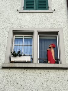 Doppelfenster an einem Wohnhaus in dessen linker Hälfte eine menschliche Skultpur mit angezogenen Beinen sitzt. Sie trägt eine rote Mütze und ein rotes Kleid
