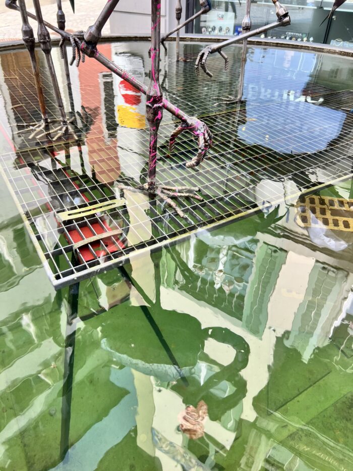 Wasserspiegelungen im Brunnen, die diversen bunten Brunnefiguren und Häuserfassaden sind auf dem grünen Wasserspiegel zu sehen