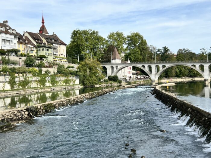 Flusskanal fließt an einer Stadt mit einer Brücke und historischen Gebäuden vorbei. Rechts und links drückt das Wasser des angrenzenden Flusses in den Kanal