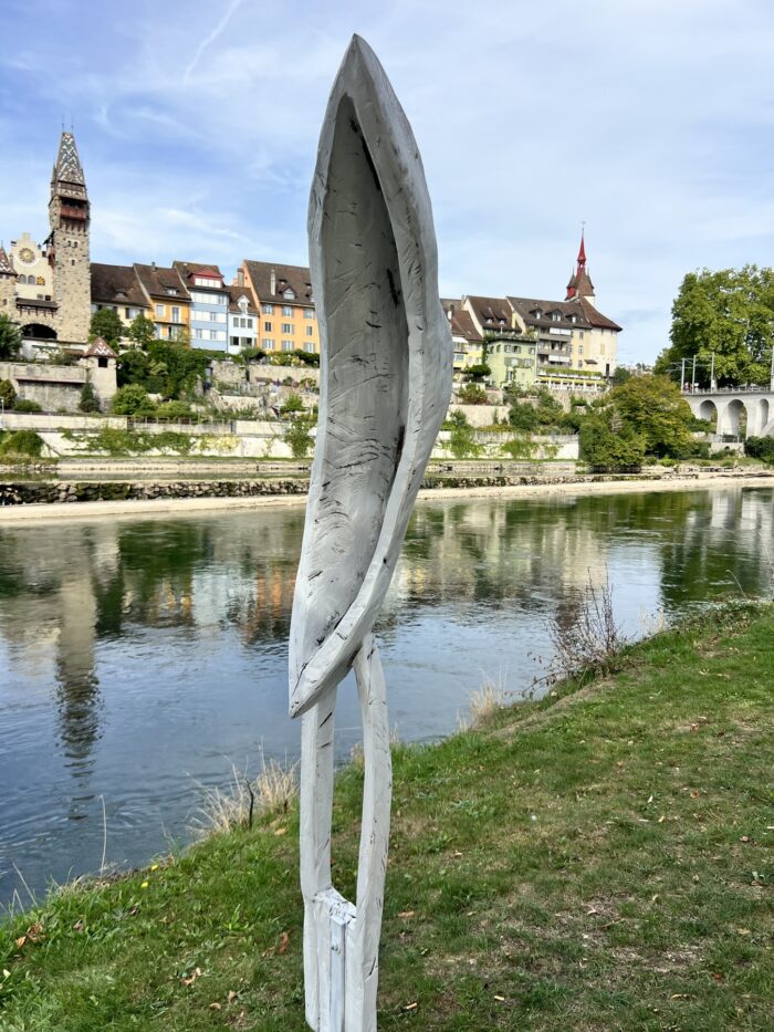 Kunstwerk aus Holz, das eine Art hochkant stehendes Boot an einer Stehle zeigt, steht auf einer Wiese am Flussufer, im Hintergrund die Stadt