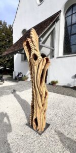 Etwa zweieinhalb Meter hohe abstrakte Holzskulptur mit rustikal oder archaisch anmutenden Formen vor Kircheneingang auf Steinplatz