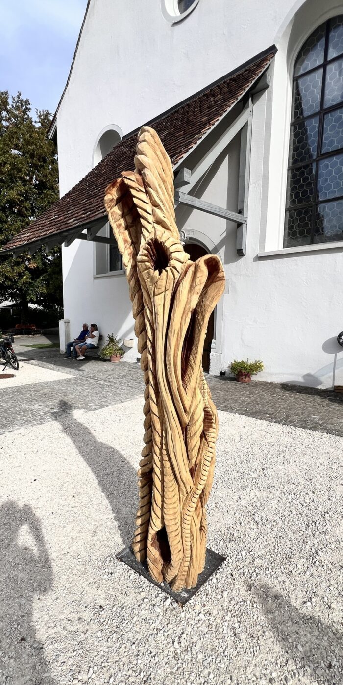 Etwa zweieinhalb Meter hohe abstrakte Holzskulptur mit rustikal oder archaisch anmutenden Formen vor Kircheneingang auf Steinplatz