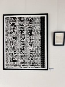 Unruhe vermittelnde Fotocollage aus vielen einzelnen schwarzweißen Aufnahnmen von Fenstern, hinter Glas gerahmt an weißer Wand, daneben in einem kleinen Rahmen ein Text, der nicht zu lesen ist
