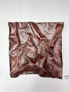 Ein mit Kupferton glasiertes Ton-Kunstwerk, das wie ein zerknittertes Stück Stoff aussieht und an einer weißen Wand hängt