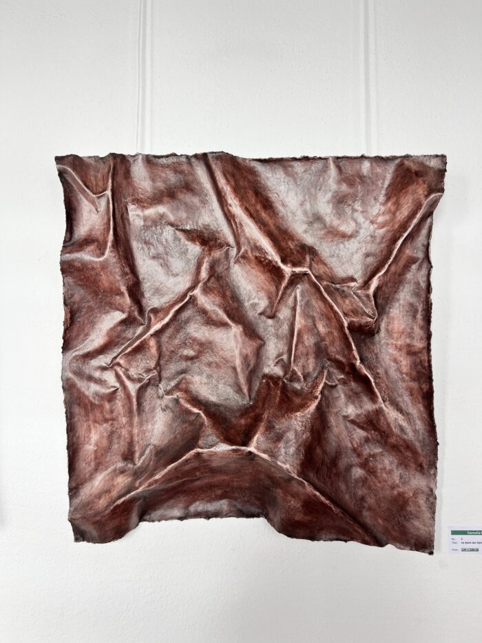 Ein mit Kupferton glasiertes Ton-Kunstwerk, das wie ein zerknittertes Stück Stoff aussieht und an einer weißen Wand hängt