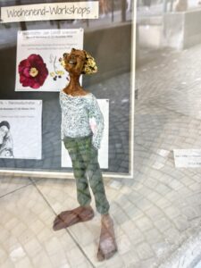 Kleine menschliche Skulptur in einem Schaufenster vor Werbung stehend, die Hände in die rückseitigen Hosentaschen gesteckt
