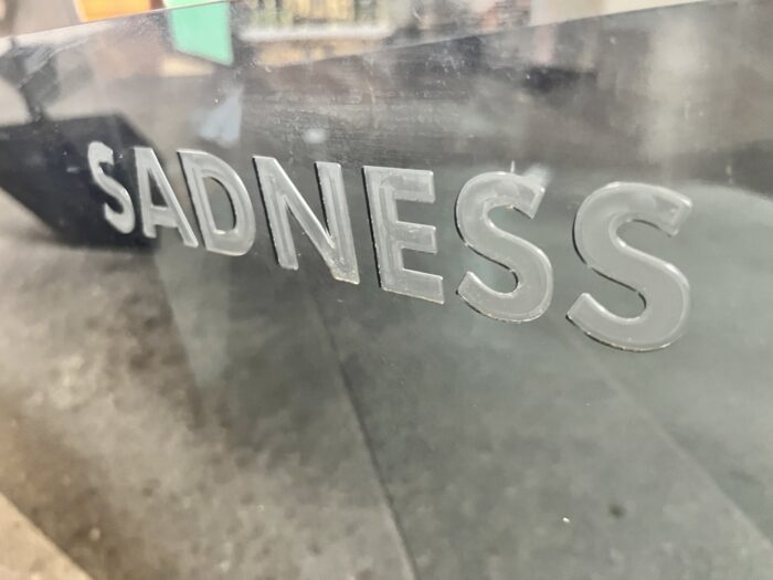 Der Schriftzug Sadness auf einer halbtransparenten graumetallenen Oberfläche, es sind auch bunte Spiegelungen aus der Umgebung zu sehen