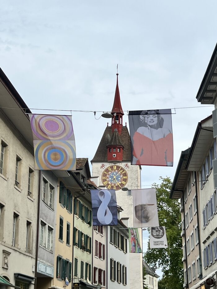 Straßenflucht mit Kirchenturm in der Mitte, davor zwei Flaggen. Die eine zeigt ein buntes, abstraktes Muster, das andere Marilyn Monroe. Weiter hinten sind weitere Flaggenpaare zu sehen