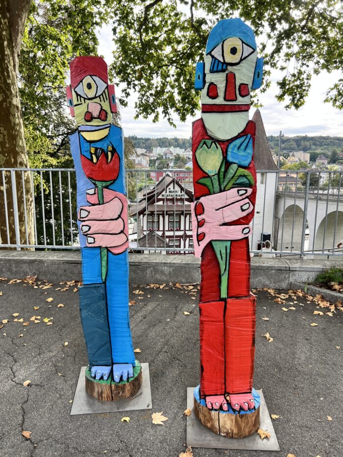 Zwei bunte, lebensgroße abstrakt-menschliche Skulpturen, die je in einer übergroßen Hand eine bunte Blumen halten. Im Hintergrund ein Zaun und dahinter tiefer unten, Hausdächer