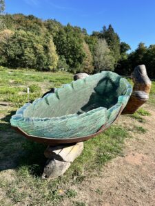 frei auf einer Wiese vor einem Waldrand stehende Skulptur: Ein Schiff aus Glasstücken zusammengeklebt, in einer Hand aus Holz.