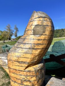 Die Daumen der Holzhand, die das Glasschiff hält, darüber Blauhimmel und im HIntergrund Wiese und Bäume.