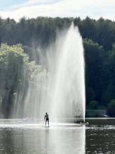 Ca. 10 Meter hohe Fontäne auf einem See. Im HIntergrund Wald am anderen Ufer. Vor der Fontäne eine kleine Gestalt auf einem Standup-Board.