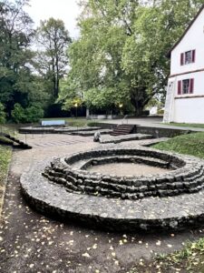 Eine archäologische Anlage bestehend aus einem kreisrunden Aufbau aus Steinen. Im Hintergrund Bäume und rechts ein Stück Hausmauer.