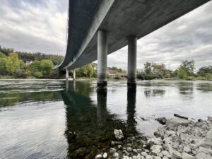Eine Betonbrücke von unten, die auf Pfeilern im Fluss steht. Vorne Kiesbank, auf der anderen Seite des Flusses sind Bäume und Häuser zu sehen.